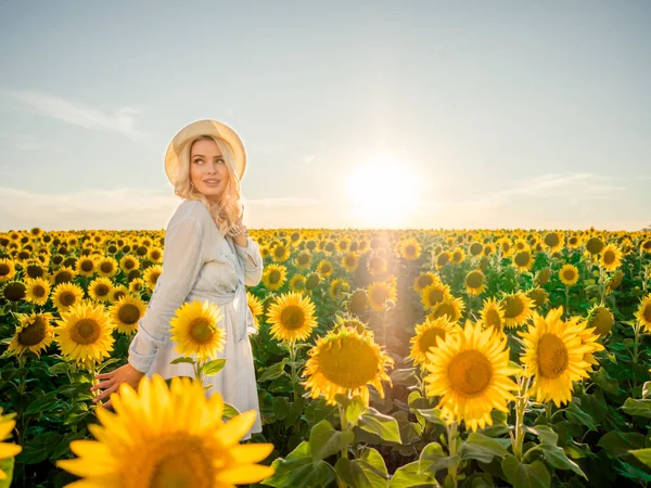 Młody piękny blond kobiety stojącej w Słonecznikowe pole. Zachód słońca na tle. Portret zmysłowy sexy dziewczyna w słomkowym kapeluszu i biały Letnia sukienka. — Zdjęcie stockowe