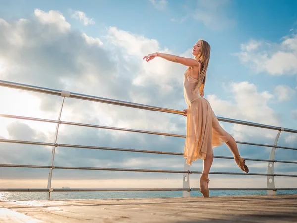 Taniec baleriny w beżowy jedwabnej sukni i pointe na nasyp powyżej plaży oceanu lub morza o świcie. Młody piękny blond kobieta z długimi włosami praktykowanie klasycznych ćwiczeń z emocjami. — Zdjęcie stockowe