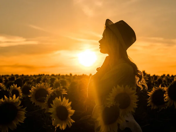 Silhouette einer jungen schönen blonden Frau, die im Sonnenblumenfeld steht. Hintergrund Sonnenuntergang. sexy sinnliches Porträt eines Mädchens mit Strohhut und weißem Sommerkleid. — Stockfoto