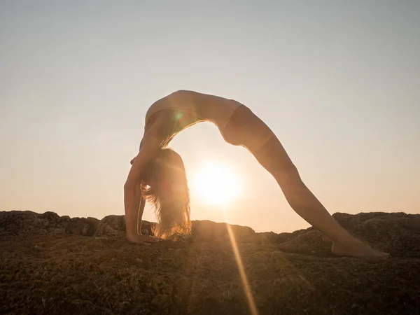 Silueta de mujer joven en traje de baño practicando yoga al amanecer increíble. Fitness, deporte, yoga y estilo de vida saludable. Chica haciendo un puente y asanas en roca en el mar . — Foto de Stock