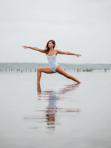 Joven mujer caucásica en traje de baño practicando yoga en agua liman, lago o río. Hermoso reflejo. Asanas complejas, equilibrio. Fitness, deporte, yoga y estilo de vida saludable . — Foto de Stock