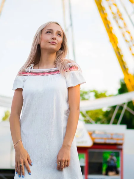 Mladá atraktivní blond dívka stojící poblíž kolotoč v zábavním parku. Portrét ženy s dlouhými vlasy. — Stock fotografie