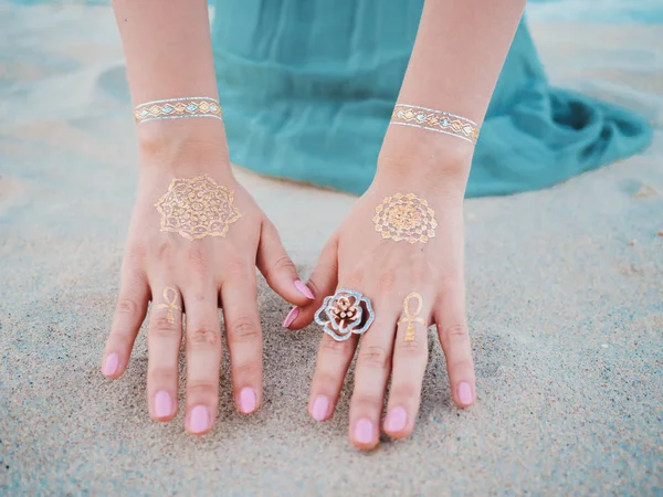 Silver and golden flash tattoo on female hands over sea or ocean background. close up hands with boho gypsy gold accessories. Woman touching sand on the beach at summer