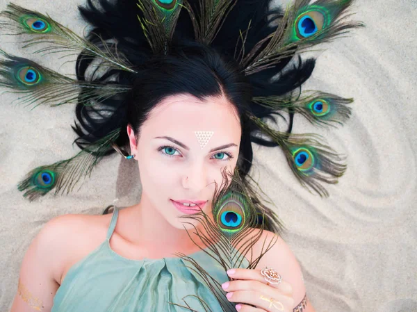 Outdoor fashion portrait of beautiful brunette lady at beach with flash tattoos lying with peacock feathers in hair. Gypsy boho style, Woman in green dress. — Stock Photo, Image