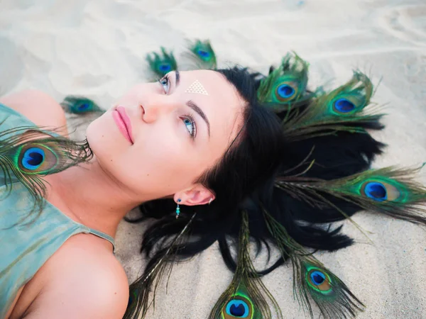 Outdoor fashion portrait of beautiful brunette lady at beach with flash tattoos lying with peacock feathers in hair. Gypsy boho style, Woman in green dress. — Stock Photo, Image