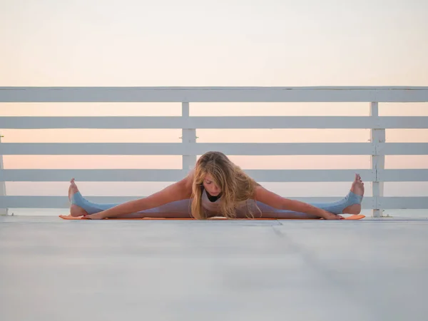 Mujer caucásica joven haciendo yoga asana en la naturaleza en un balcón de madera o puente con vistas al mar. Mujer practicando en el océano relajándose en la naturaleza. Chica en ropa deportiva . — Foto de Stock