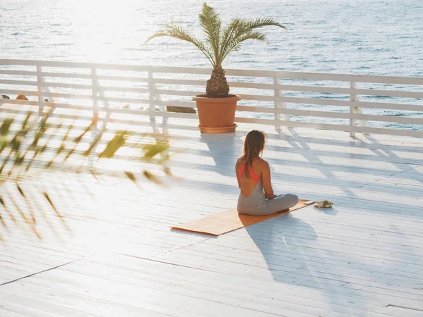 Giovane donna caucasica che fa yoga asana nella natura su un balcone in legno con vista sul mare. Donna che fa pratica sull'oceano rilassandosi nella natura. Meditazione. Ragazza in abbigliamento sportivo . — Foto Stock