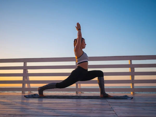 Молода біла жінка робить йогу асану в природі на дерев'яному балконі або мосту з видом на море. Жінка практикує в океані, розслабляючись в природі. Дівчина в спортивному одязі . — стокове фото