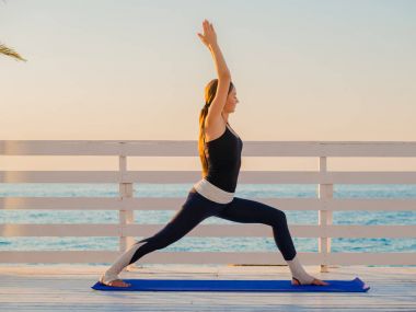 Bir ahşap balkon ya da denize bakan köprü doğada Yoga asana yaparken genç beyaz kadın. Kadının doğada rahatlatıcı okyanus üzerinde pratik yapması. Kız spor giyim.