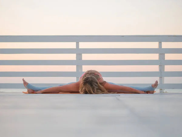 Mujer caucásica joven haciendo yoga asana en la naturaleza en un balcón de madera o puente con vistas al mar. Mujer practicando en el océano relajándose en la naturaleza. Chica en ropa deportiva . — Foto de Stock