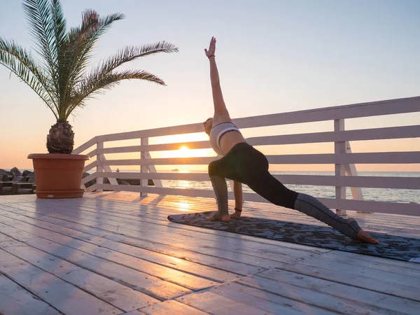Giovane donna caucasica che fa yoga asana nella natura su un balcone in legno o ponte con vista sul mare. Donna che fa pratica sull'oceano rilassandosi nella natura. Ragazza in abbigliamento sportivo . — Foto Stock