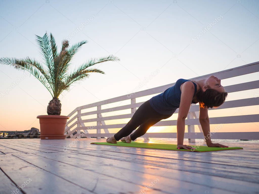 Young caucasian woman doing yoga asana in the nature on a wooden balcony or bridge overlooking sea. Woman doing practice on the ocean relaxing in nature. Girl in sports wear.