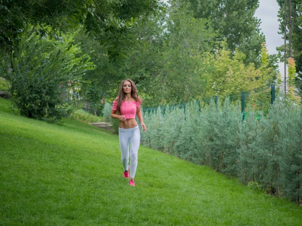 Vacker ung kaukasisk kvinna i fitnesscenter slitage arbetande ute i en park. Kvinna med långt hår och sexig outfit gör sport övningar, kör i skogen på sommaren. — Stockfoto