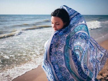 Gypsy young brunette girl wearing white maxi long dress standing near sea or ocean with mandala silk scarf in hands. Bohemian clothing style. Boho lifestyle.