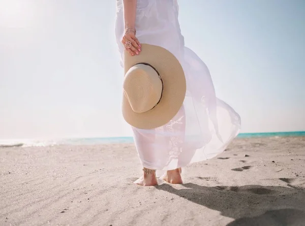 Piękna dziewczyna kaukaski trzyma kapelusz na brzegu morza. Kobieta ubrana w długiej, białej sukni, stały na piasku. Wietrznej pogody. Koncepcja podróży. — Zdjęcie stockowe