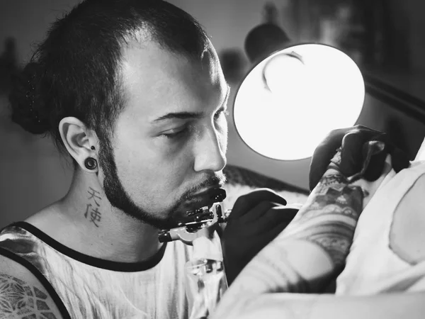 Portrait of man tattoo master with dreadlocks showing process of creation tattoo on female body under the lamp light.Professional artist working in salon, close up view.