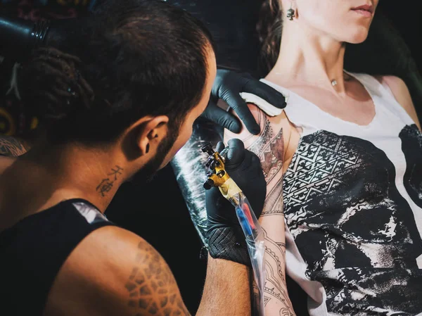 Portrait of man tattoo master with dreadlocks showing process of creation tattoo on female body under the lamp light.Professional artist working in salon, close up view.