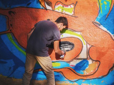 Yaratıcı sanat - genç çocuk sokak duvarına sprey spreyle renkli soyut süsleme grafitisi çiziyor. Arka plan, genel plan
