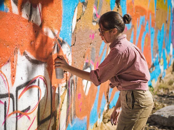 Beautiful Young Girl making a colorful graffiti with aerosol spray on urban street wall. Cinematic toned shot. Creative art. Talented woman in glasses drawing picture. — Stock Photo, Image