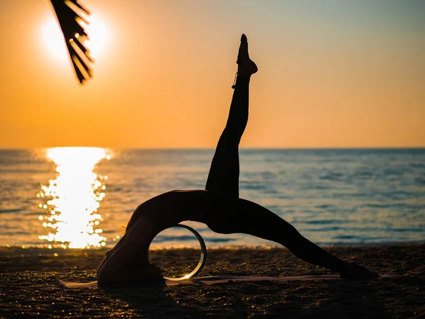 Силуэт красивой молодой женщины, практикующей утреннюю йогу на песчаном пляже с колесом для йоги . — стоковое фото