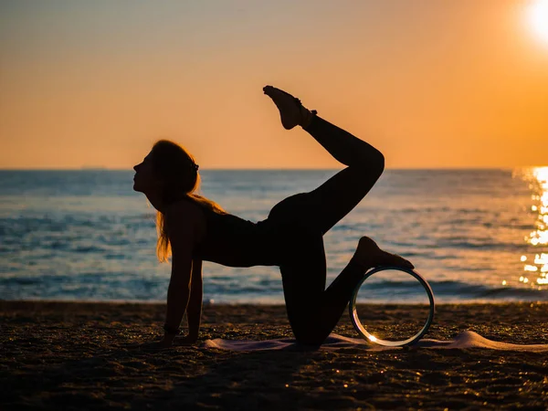 Silhouette di bella giovane donna che pratica yoga mattutino sulla spiaggia di sabbia con ruota yoga . — Foto Stock