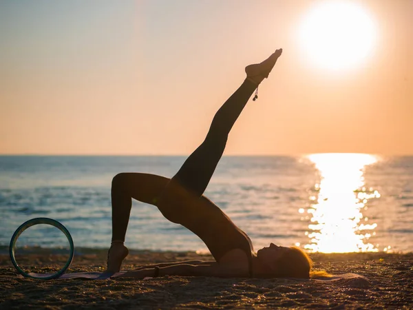 Silhouette di bella giovane donna che pratica yoga mattutino sulla spiaggia di sabbia con ruota yoga . — Foto Stock