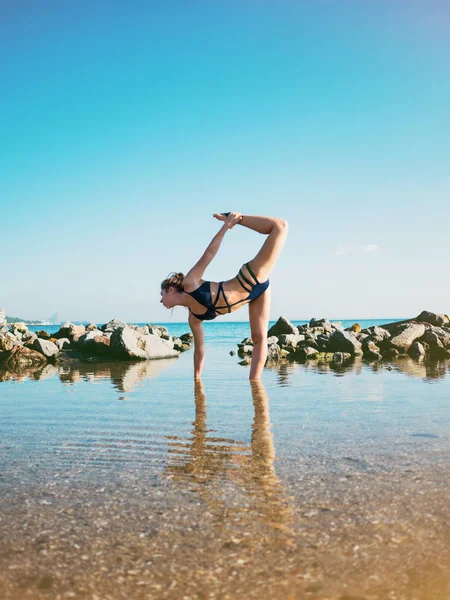 Giovane donna caucasica in costume da bagno praticare yoga in acqua di mare, lago o fiume. Bel riflesso. Asana complesse, equilibrio. Fitness, sport, yoga e stile di vita sano concetto . — Foto Stock