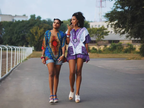 Two happy beautiful afro-american women friends walking outdoor. Young mixed race girls wearing colorful clothing communicates, enjoys the meeting and laughing — Stock Photo, Image