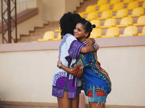Zwei glückliche schöne afroamerikanische Freundinnen, die sich im Freien umarmen. junge Mischlingsmädchen in bunten Kleidern kommunizieren, genießen das Treffen und die Begrüßung — Stockfoto