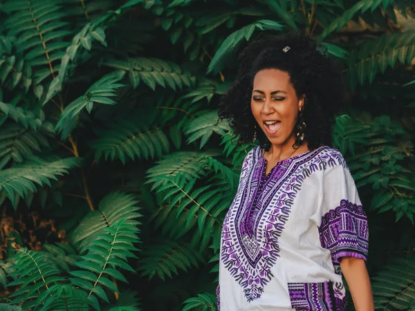 Afro-american woman singing outdoor. Multi ethnic girl wearing colorful clothing posing, enjoys the nature. Green tropical background. Success concept — Stock Photo, Image