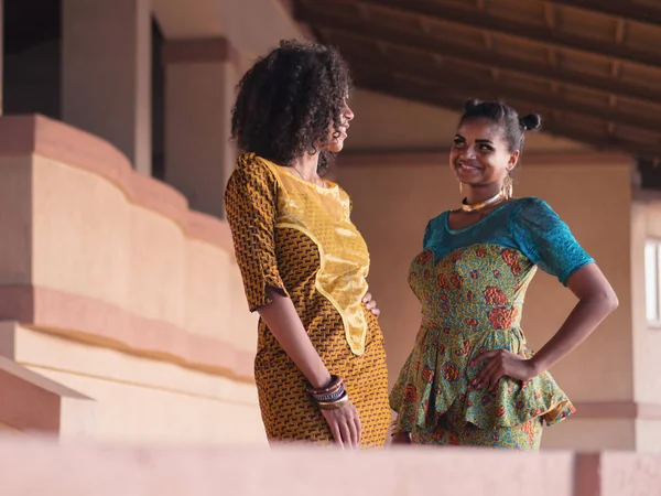 Two happy beautiful afro-american women friends outdoor. Multi ethnic girls wearing african long dresses communicates, enjoys the meeting and laughing — Stock Photo, Image