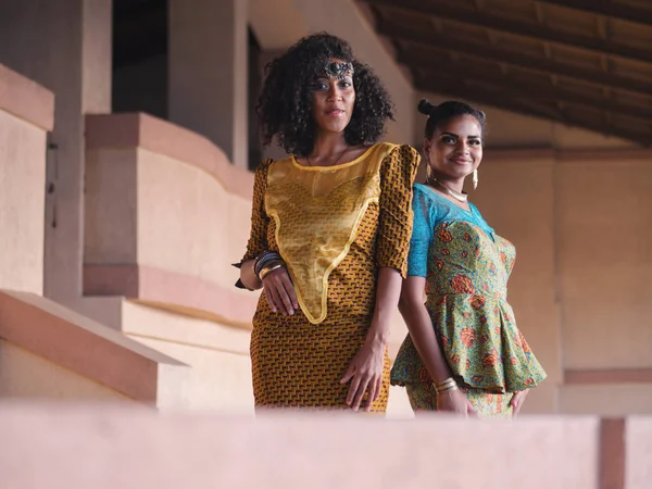 Two happy beautiful afro-american women friends outdoor. Multi ethnic girls wearing african long dresses communicates, enjoys the meeting and laughing — Stock Photo, Image