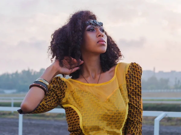 Portrait of young afro-american gypsy woman in colorful yellow traditional dress and silver crown on forehead smiling and dancing close to camera. Sexy fashion girl with curly hair — Stock Photo, Image