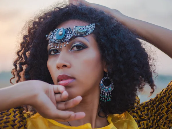 Portrait of young afro-american gypsy woman in colorful yellow traditional dress and silver crown on forehead smiling and dancing close to camera. Sexy fashion girl with curly hair — Stock Photo, Image