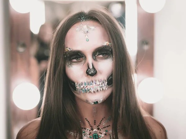Korkunç genç kız soyunma odasında yaratıcı Cadılar Bayramı savaş-boya ile. Göz alıcı kafatası ile rhinestones ve payetler portresi. Kutlama için profesyonel makyaj.
