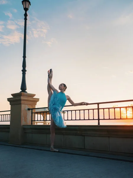 Dancing ballerina in blue ballet tutu and point on embankment above ocean or sea at sunset. Young beautiful brunette woman practicing stretching and exercises.Copy space — Stock Photo, Image