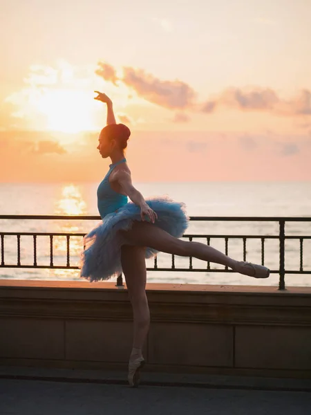 Dansende ballerina in blauwe ballet tutu en punt op talud boven de oceaan of de zee bij zonsondergang. Jonge mooie brunette vrouw beoefenen van rekken en oefeningen. — Stockfoto