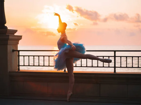 Dansende ballerina in blauwe ballet tutu en punt op talud boven de oceaan of de zee bij zonsondergang. Jonge mooie brunette vrouw beoefenen van rekken en oefeningen. — Stockfoto