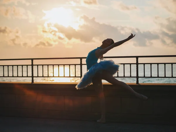 Ballerina danzante in tutù blu balletto e punto su argine sopra l'oceano o mare al tramonto. Giovane bella donna bruna che pratica stretching ed esercizi. — Foto Stock