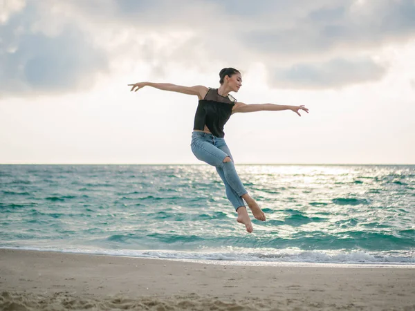 Mulher jovem em estilo casual - ganga e top preto fazendo balé na praia. Bailarina atraente pratica em saltar na costa arenosa no outono. Espaço de cópia — Fotografia de Stock