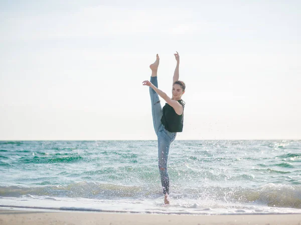 Bela cena de uma bailarina dançante em top preto e jeans no oceano de praia arenosa ou mar de manhã. Mulher morena bonita praticando alongamento e exercícios.Espaço de cópia — Fotografia de Stock