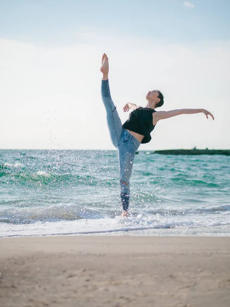 Bela cena de uma bailarina dançante em top preto e jeans no oceano de praia arenosa ou mar de manhã. Mulher morena bonita praticando alongamento e exercícios.Espaço de cópia — Fotografia de Stock