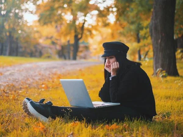 Sad young woman using laptop outdoor sitting on yellow grass. Girl using digital tablet pc in the autumn park. Trendy student in fashion hat using tablet after school.