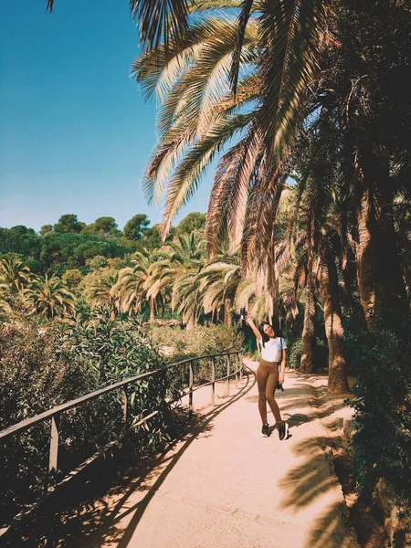 年轻的时尚美丽的晒黑的女人与黑头发走在巴塞罗那公园盖尔热带棕榈树之间的路径。旅游理念. — 图库照片