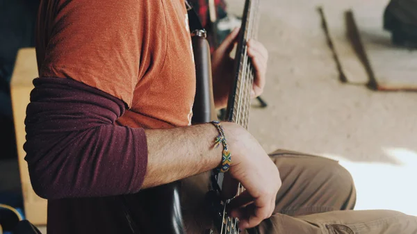 Hombre tocando el bajo en el concierto en la calle. Vista de cerca de guitarrista toca en instrumento musical de madera marrón. Músico callejero con un brazalete hippie de mimbre en su brazo . — Foto de Stock