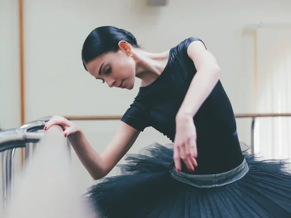 Ballerina in een zwarte tutu en pointe strekt zich uit over barre in ballet sportschool. Vrouw stond in de buurt van bar en spiegel, perfomance voorbereiden. — Stockfoto