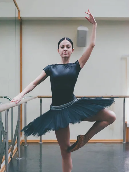 Ballerina in een zwarte tutu en pointe strekt zich uit over barre in ballet sportschool. Vrouw stond in de buurt van bar en spiegel, perfomance voorbereiden. — Stockfoto