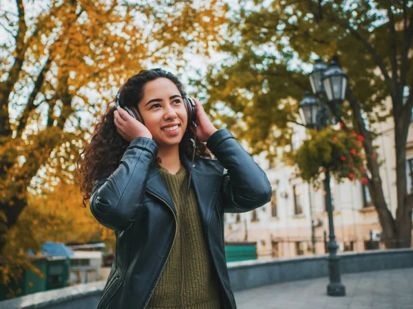 Łacińska kręcone włosy dziewczyna słuchania muzyki w słuchawkach w jesienny park. Piękna panienka hiszpanin uśmiechający się, śpiewać i cieszyć się melodii odkryty. — Zdjęcie stockowe