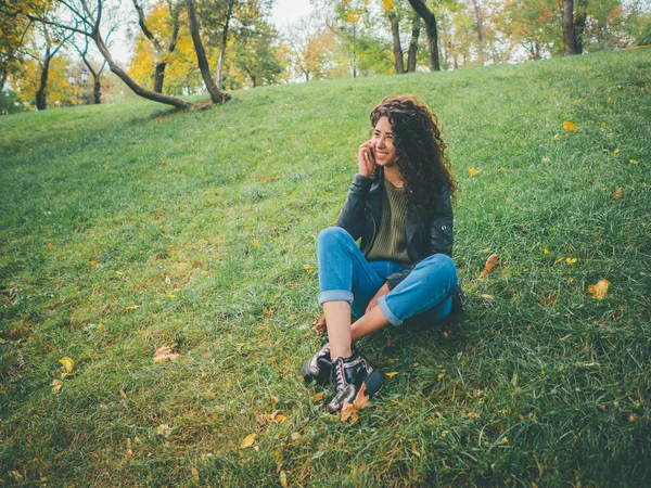 Piękne młode kręcone włosy Łacińskiej dziewczyna za pomocą jej telefon komórkowy, odkryty. Portret zrelaksowany hiszpanin panienka w zielonym parku, rozmowy na telefon. — Zdjęcie stockowe