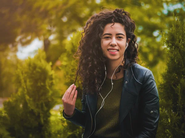 Dość młoda dziewczyna Łacińskiej czarne kręcone włosy długie słuchanie muzyki z słuchawki i smartfona lub odtwarzacza na opuszczony jesienią ulicy. Kobieta korzystających chwilę. — Zdjęcie stockowe